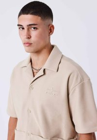 Polo shirt beige a maniche corte con tessuto testurizzato, dotato di colletto, chiusura con bottoni e logo ricamato sul lato sinistro.
