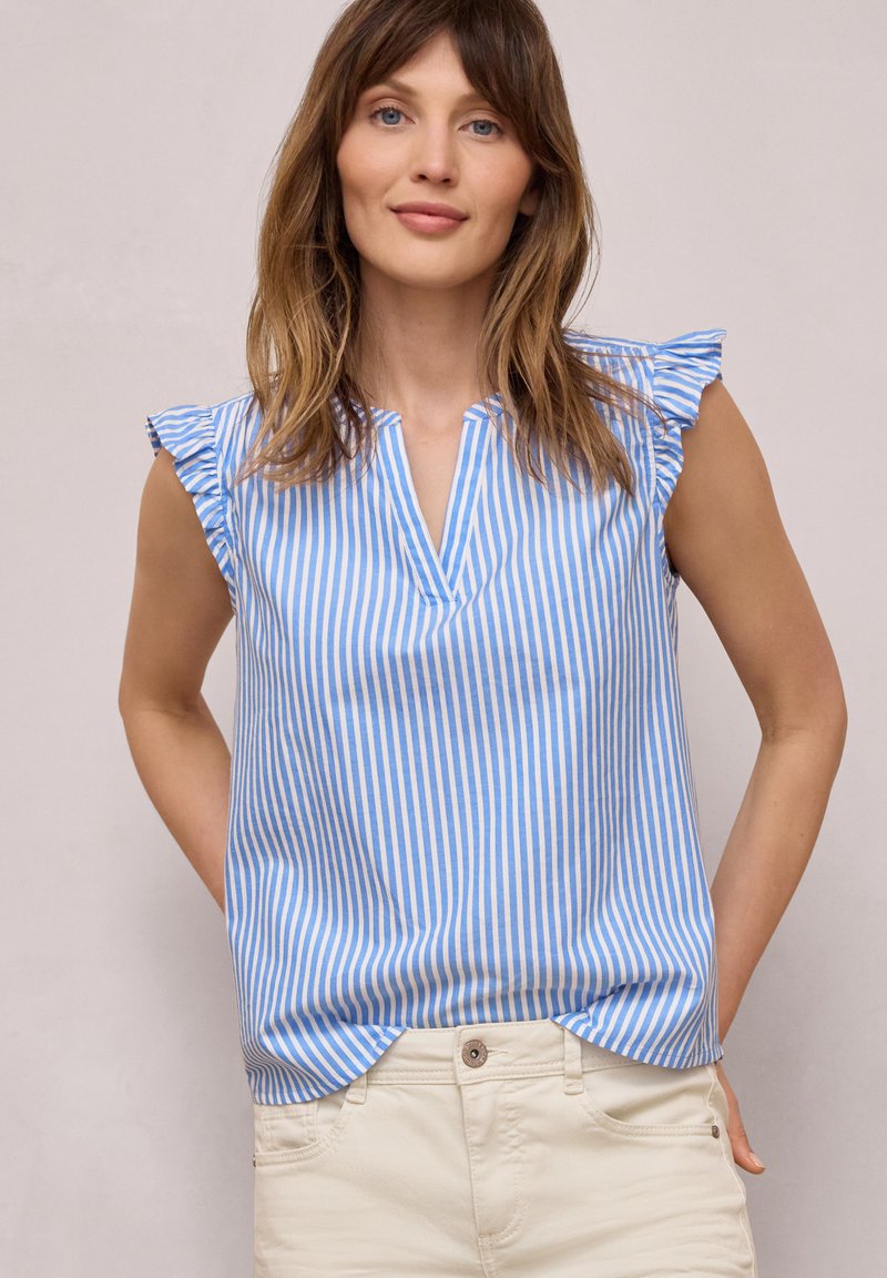 Mujer que lleva una blusa sin mangas de rayas azules y blancas con volantes en los hombros y pantalones beige claro, de pie frente a un fondo liso.