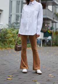 Person som bär en vit oversized knappad skjorta, bruna utsvängda byxor, vita sneakers och håller en liten handväska med ormskinnsmönster utomhus.