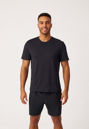 Man staat en lacht, draagt een effen zwart T-shirt met korte mouwen en zwarte korte broek tegen een neutrale achtergrond.