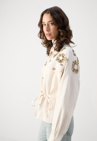 Camicia bianca con bottoni e ricami floreali dorati, vita con coulisse e maniche lunghe a sbuffo. Tessuto texturizzato, vestibilità casual.
