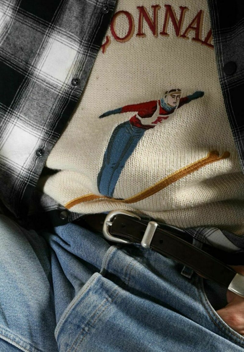 Maglione di lana con un design di uno sciatore ricamato in rosso, bianco e blu, indossato sotto una camicia a scacchi bianca e nera, abbinato a jeans di denim.