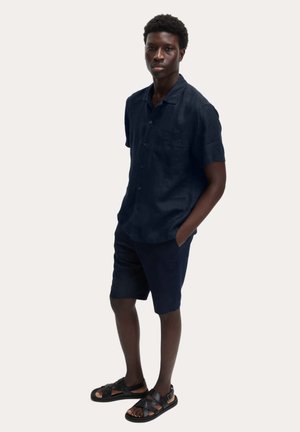 Marineblauw linnen overhemd met korte mouwen en een borstzak, gecombineerd met bijpassende korte broek en zwarte sandalen met bandjes, afgebeeld tegen een lichte achtergrond.