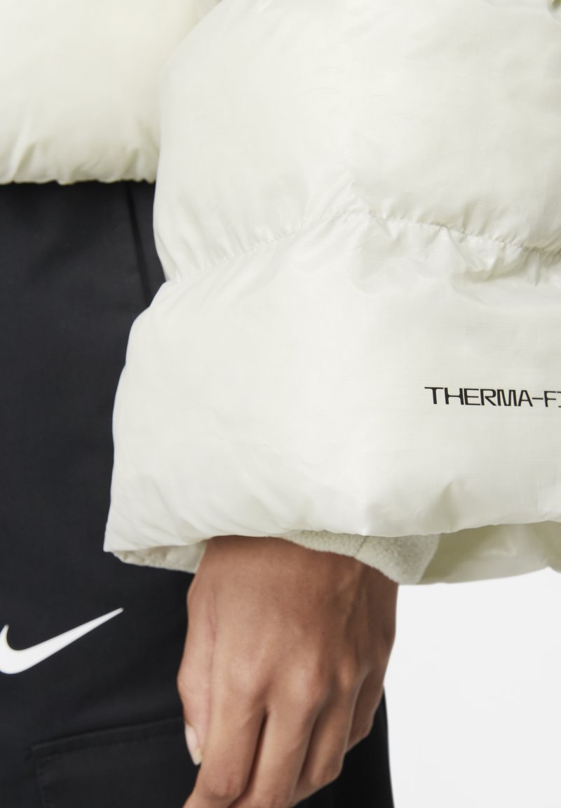Chaqueta blanca acolchada con un suave forro interior y puños elásticos. Logo "THERMA-FIT" impreso en negro en la manga, combinada con pantalones oscuros.