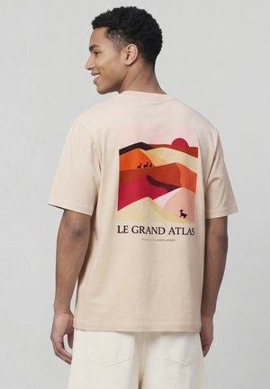 Homme de dos portant une chemise beige avec un paysage désertique multicolore, des animaux et le texte « Le Grand Atlas, Maroc, Afrique du Nord » dans le dos.