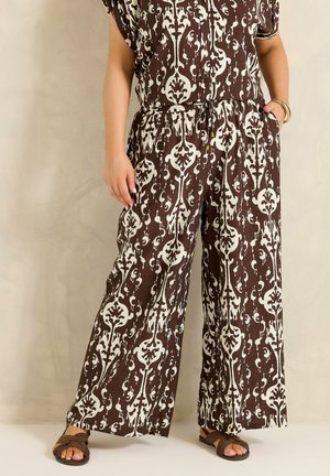 Femme portant un pantalon large marron et blanc à motifs, des sandales marron et un haut assorti à manches courtes, debout avec une main dans la poche.