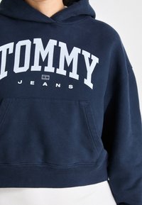 Felpa crop con cappuccio blu navy in cotone, con grande logo bianco "TOMMY" e tasca frontale. Tessuto liscio con polsini a costine.