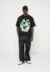 Tricou grafic negru cu imprimeu de craniu și flăcări verzi, asortat cu pantaloni largi negri lucioși și teniși albi cu dungi negre.