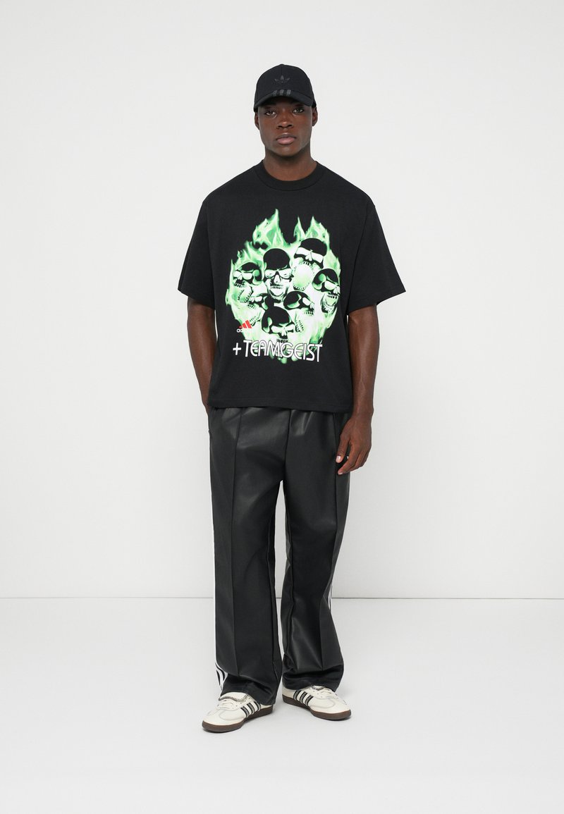Camiseta gráfica negra con estampado de calavera y llamas verdes, combinada con pantalones anchos negros brillantes y zapatillas blancas con rayas negras.