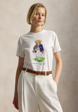 Femeie cu păr scurt și blond, purtând un tricou alb cu un urs îmbrăcat într-un sacou care ține rachete de tenis, asortat cu pantaloni albi și o curea maro.