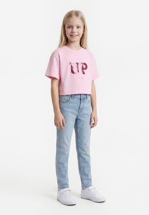 Ung pige med langt blondt hår iført en pink croppet T-shirt med "UP" i pailletter, lyseblå jeans og hvide sneakers, smilende.