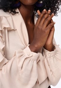 Beżowa bluzka z falbankami i bufkami. Miękka tekstura z zapięciami na guziki w mankietach. Ręce złożone przed sobą.