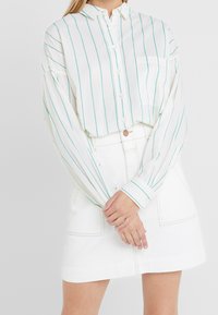 Camicia bianca con bottoni, caratterizzata da righe verticali turchesi, maniche lunghe e una tasca frontale, abbinata a una gonna di jeans bianca.