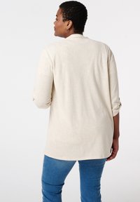 Chemise beige à manches longues avec un ourlet arrondi et des manches retroussées, associée à un jean bleu ajusté. Tissu lisse avec une texture subtile.
