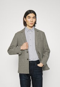 Blazer cu model houndstooth în nuanțe de maro, cu două butoane, asociat cu o cămașă gri deschis și blugi de culoare albastru închis, subliniind o croială structurată.