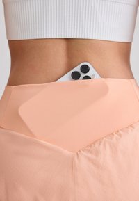 Smartphone partiellement glissé dans la ceinture arrière d'un pantalon taille haute couleur pêche porté par une personne en haut court blanc.