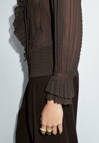 Blouse marron plissée en tissu transparent, avec des accents froncés aux poignets et un corsage ajusté. Présente des textures superposées et une coupe douce.