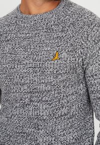 Szary sweter z dzianiny z teksturowanym wzorem i żółtym haftowanym logo ptaka po lewej stronie klatki piersiowej. Okrągły dekolt i długie rękawy.
