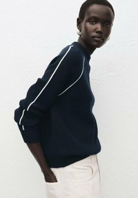 Marineblauer Pullover mit gerippter Struktur, weißen Paspeln entlang der Schultern und lockerem Schnitt, kombiniert mit hellfarbigen Hosen.