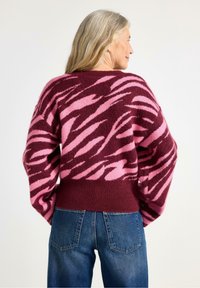 Maglione peloso con sfondo color bordeaux e audaci strisce zebrate rosa, vestibilità oversize e orlo a coste. Abbinato a jeans in denim blu.
