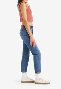 Femme portant un jean bleu taille haute, des baskets blanches avec semelles en gomme, des chaussettes blanches et un crop top sans manches couleur rouille avec des boutons.