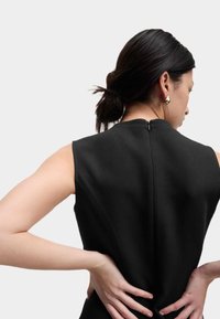 Schwarzes ärmelloses Kleid mit hohem Halsauschnitt, strukturiertem Stoff, subtilen Nähdetails und einem Reißverschluss hinten, von hinten betrachtet.