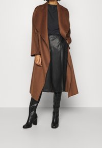 Cappotto di lana marrone con davanti aperto, indossato sopra una camicia nera testurizzata e una gonna di pelle plissettata nera, abbinato a stivali neri al ginocchio.