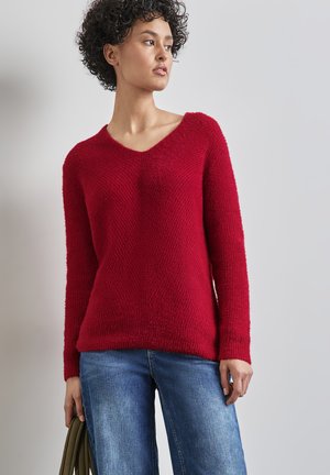 Rød strikket sweater med V-hals, struktureret stof, lange ærmer og en afslappet pasform. Stylingset med blå bredbenede jeans og en grøn taske.