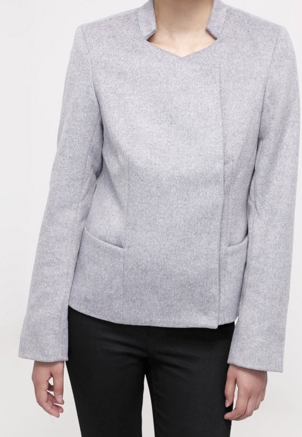 Femme portant une veste gris clair à manches longues avec un col unique et un pantalon noir, debout devant un fond uni.