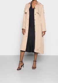 Beige lång trenchcoat med knäppning och klaffdetalj, lager-på-lager över en svart midiklänning, kombinerad med svarta sandaler med remmar.