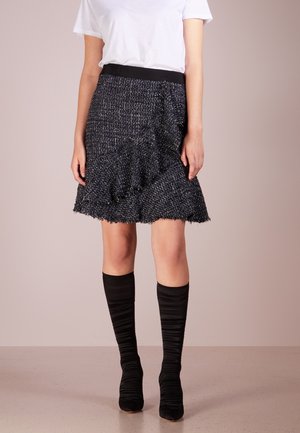 Kobieta ubrana w teksturowaną, czarno-białą spódnicę z grubym wzorem i postrzępionym brzegiem, białą krótką koszulę oraz czarne marszczone kozaki za kolano.