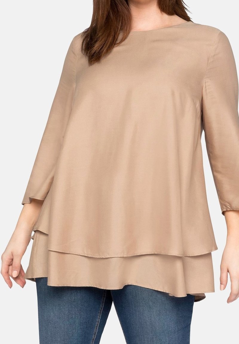 Blouse beige à plusieurs couches avec une texture lisse, col rond et manches trois-quarts, associée à un jean en denim bleu.