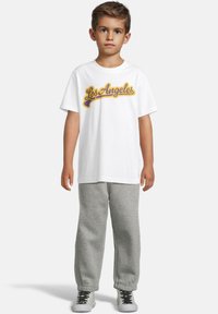 Gutt som står med en hvit "Los Angeles" t-skjorte, grå sweatpants og hvite sneakers med svarte lisser på enkel lys bakgrunn.