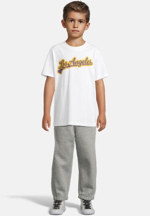 Gutt som står med en hvit "Los Angeles" t-skjorte, grå sweatpants og hvite sneakers med svarte lisser på enkel lys bakgrunn.