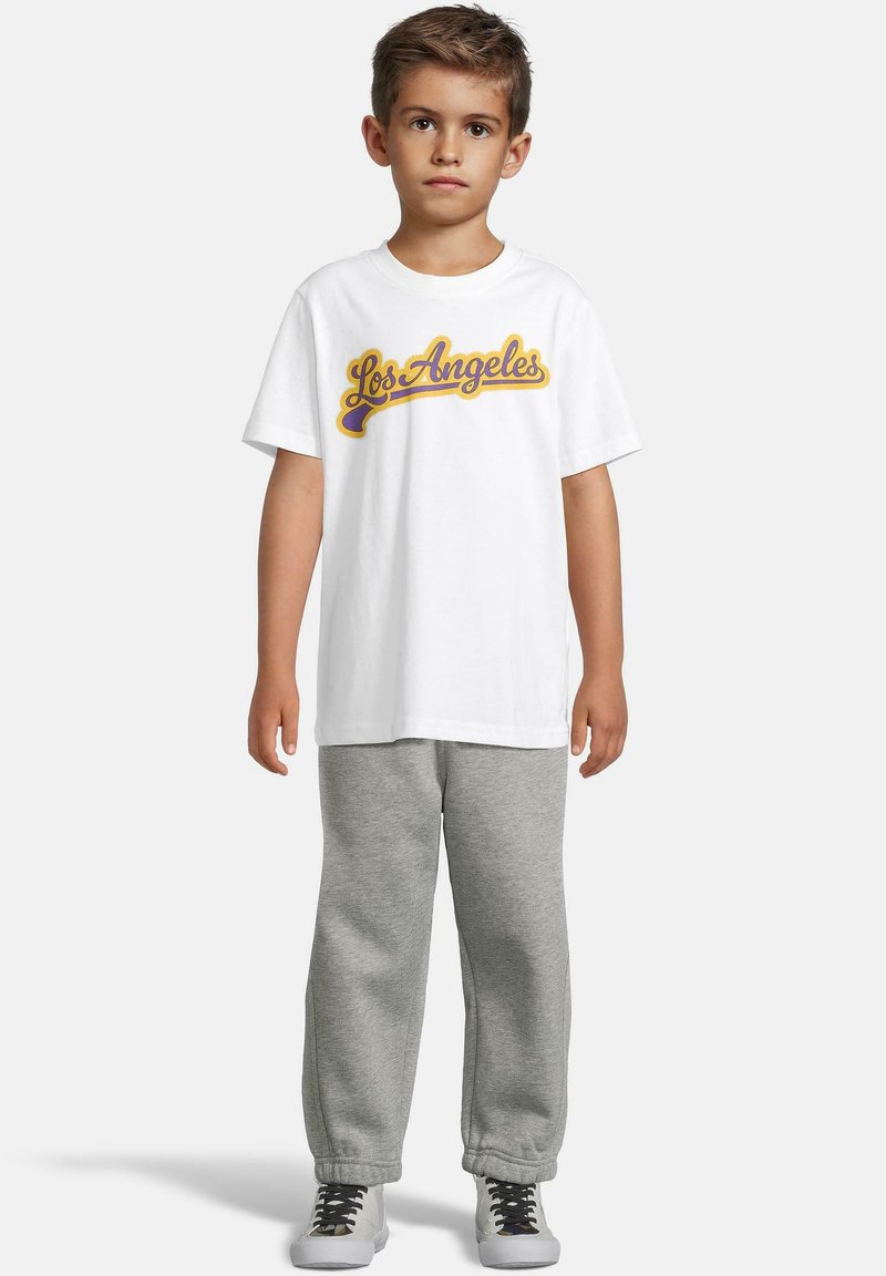 Gutt som står med en hvit "Los Angeles" t-skjorte, grå sweatpants og hvite sneakers med svarte lisser på enkel lys bakgrunn.