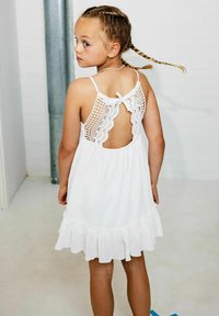 Weißes ärmelloses Kleid mit Spitzenrücken, gerafftem Saum und überkreuzten Trägern. Weicher Stoff und detaillierte Häkelakzente.