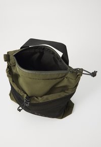 Mala u boji maslina s zatvaračem na zip, ima mrežastu džep i crnu traku za nošenje. Tkanina ima glatku, mat površinu.