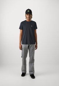 Dunkelgraues Baumwoll-T-Shirt mit kleinem Logo, kombiniert mit hellgrauen, stark ausgewaschenen Jeans mit Seitentaschen und fransigen Kanten. Schwarze Mütze und Schuhe.