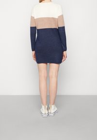 Robe-pull à manches longues avec des rayures crème, beige et bleu marine. Texture en maille douce, coupe ajustée, longueur mi-cuisse. Portée avec des baskets montantes blanches.