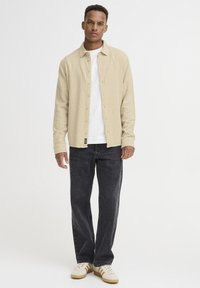Beige ribbet skjorte lagdelt over en hvid t-shirt, parret med mørk denim jeans og creme sneakers. Enkel knaplukning, afslappet pasform.