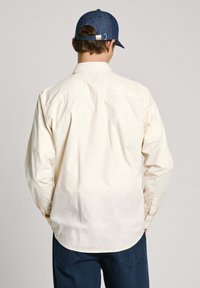 Camisa de manga larga color crema hecha de algodón, con un dobladillo curvado y puños con botones. Lleva puesta una gorra de mezclilla oscura.