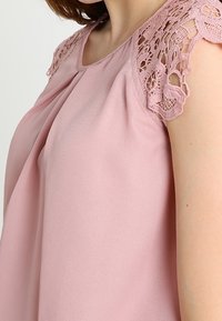 Blusa rosa caratterizzata da un tessuto morbido, design anteriore plissettato e dettagli in pizzo intricati sulle spalle per un tocco di texture e interesse visivo.