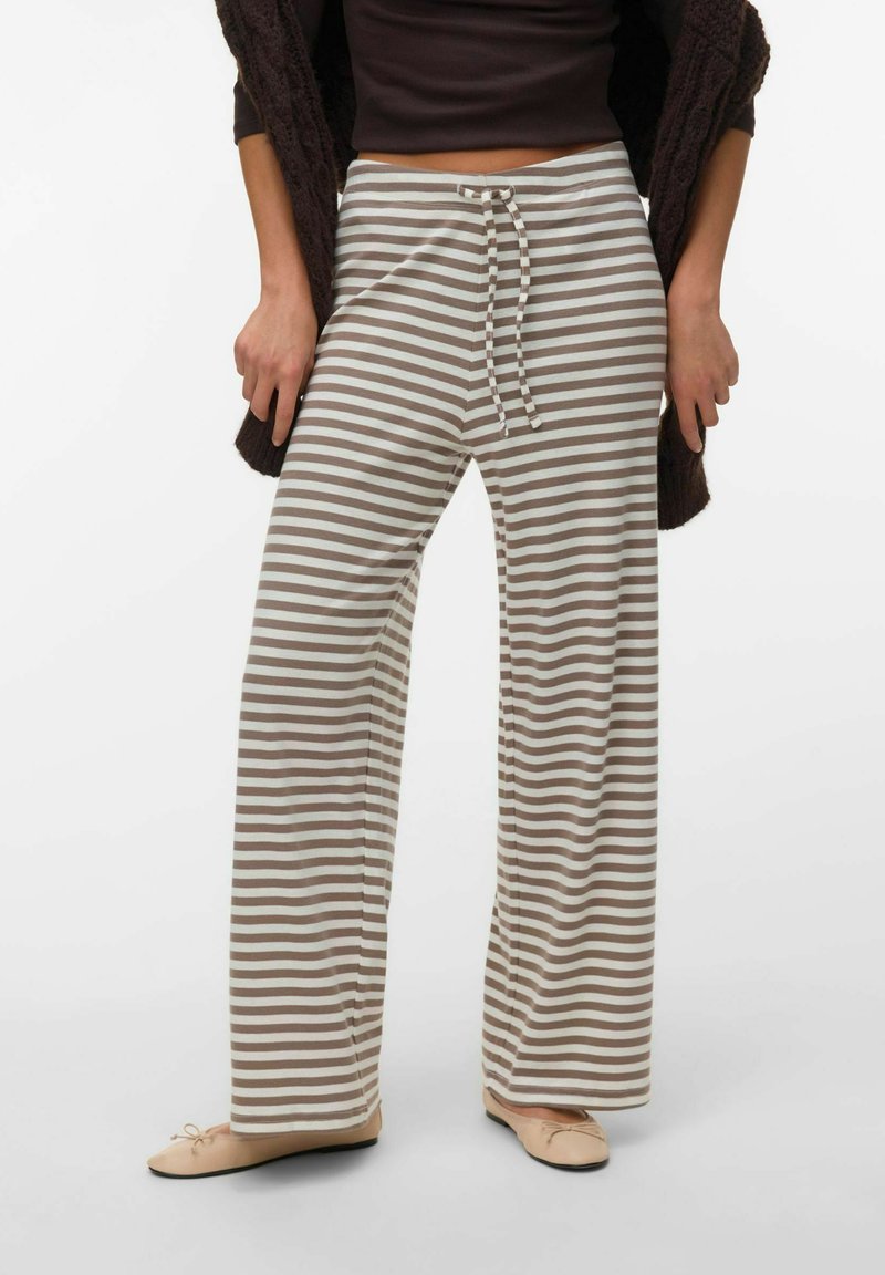 Personne portant un pantalon large à rayures horizontales beige et blanc avec cordon de serrage, un haut noir et des ballerines beige sur un fond uni.