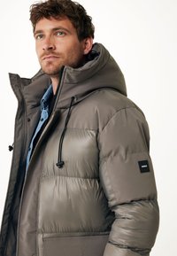 Giacca imbottita verde olivastro con colletto alto. Presenta un cappuccio regolabile e una patch con logo sulla manica sinistra. Texture liscia, leggera.