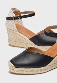 Sandalias de cuña de cuero negro con parte trasera de tela crema texturizada, suela envuelta en yute y una correa ajustable en el tobillo para un ajuste seguro.