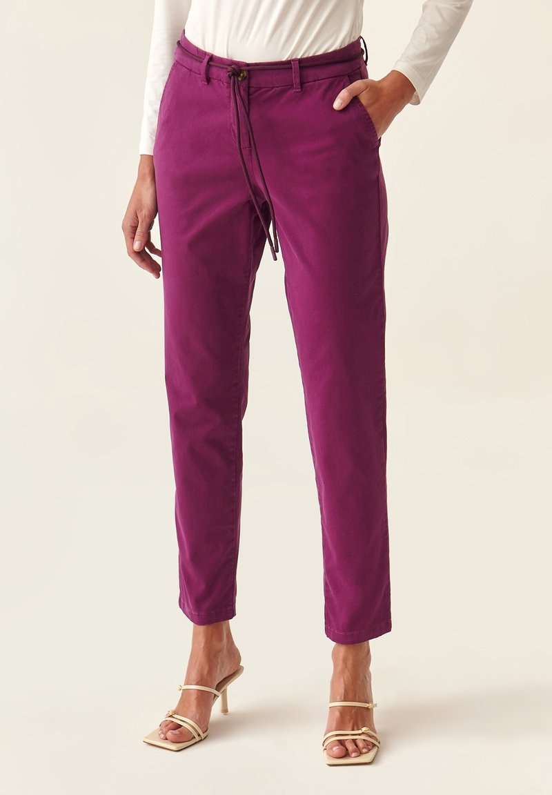 Femme portant un pantalon fuselé violet avec un lien à la taille et des sandales à talons beiges, debout avec une main dans la poche sur un fond neutre.