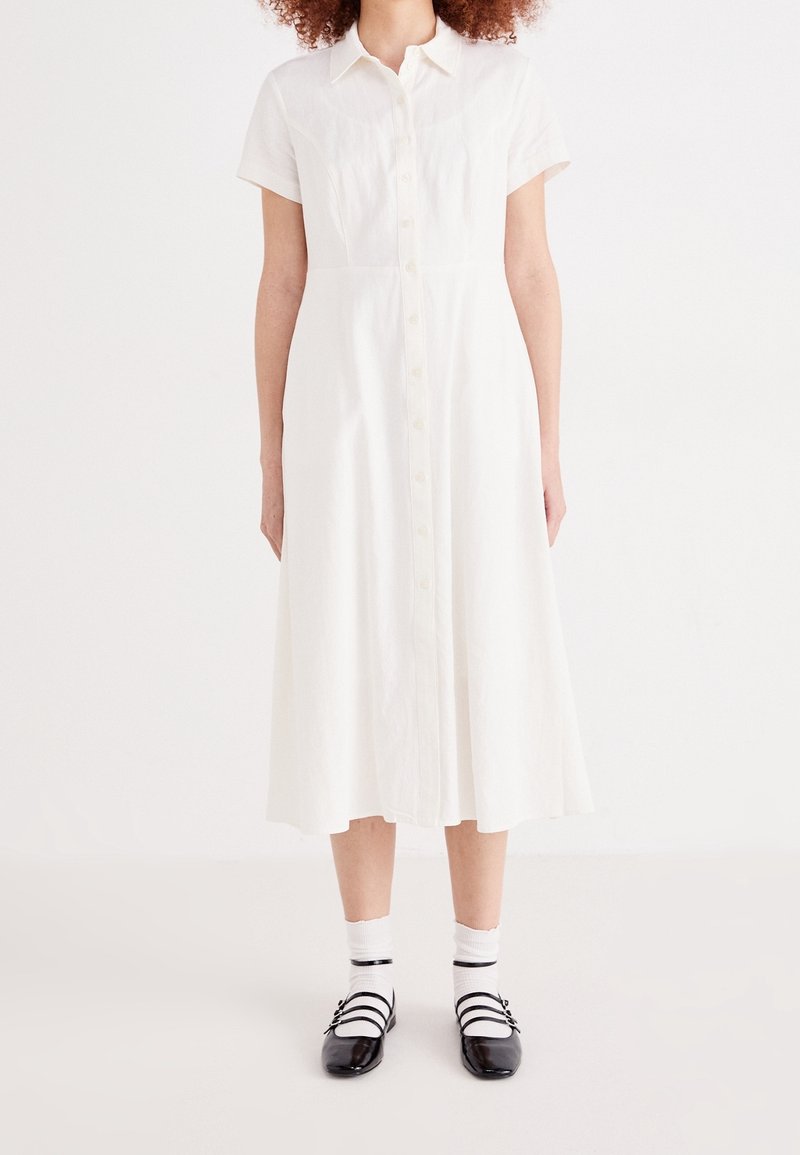 Femme portant une robe midi blanche à manches courtes avec boutons, des chaussettes blanches et des chaussures Mary Jane noires, debout sur un fond uni.
