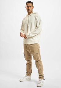 Cream hoodie with a logo, khaki cargo pants featuring multiple pockets, and white sneakers with black accents. Smooth fabric and relaxed fit.