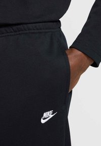 Melni sporta džinsi ar mīkstu tekstūru, ar baltu Nike logo uz augšstilba un sānu kabatu. Ikdienas dizains.