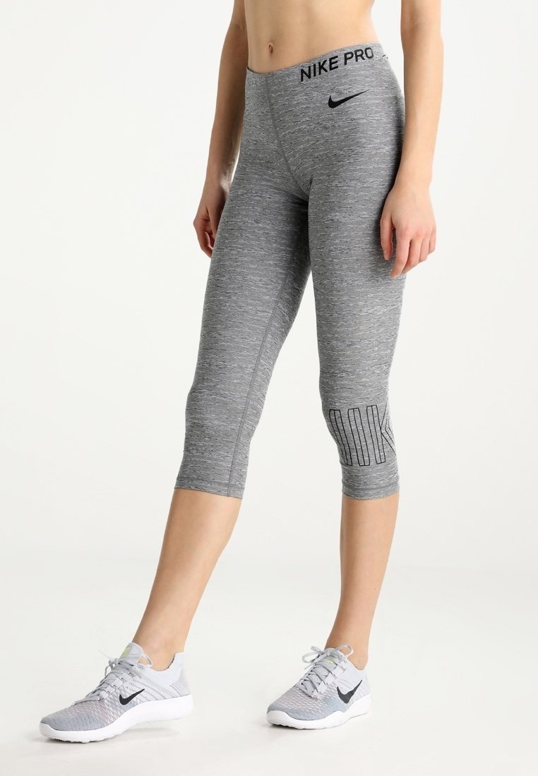 Femme portant des leggings Nike Pro gris courts et des baskets Nike grises assorties sur un fond blanc uni.